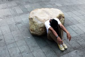 Dieses Bild zeigt einen Mann der vor einem großen Stein sitzt. Er lässt seinen Kopf zwischen seinen Beinen hängen und wirkt erschöpft. Dieses Bild soll einen Menschen mit einer psychischen Erkrankung symbolisieren.